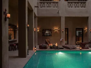 A modern residential lounge area with a poolside setup, featuring concrete walls and floors, soft furnishings, and warm light