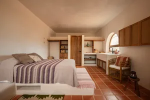 A minimalist bedroom with terracotta tiles features a bed dressed in striped textiles, a wicker chair with striped cushions,