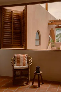 Minimalist residential balcony featuring woven rattan chair, terracotta tiles, and wooden shutters.