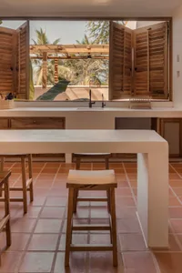 Modern minimalist kitchen with white countertops, wooden stools, and open shutters showcasing a view of palm trees and clear 