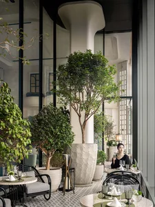 Modern restaurant interior with concrete columns, potted plants, and tables set with plates and glasses.