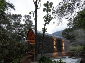 A modern stone house exterior at night with a small pool in front and a tree nearby.