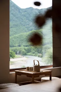 A minimalist teahouse interior featuring a bamboo table with a teapot and cups, set against a large window overlooking lush g