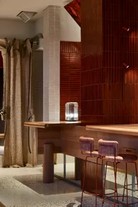 Modern coffee shop interior with wooden counter and metal stools, featuring red tiles and textured curtains.