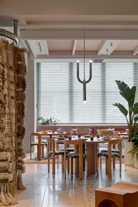 A modern restaurant dining room featuring wooden tables and chairs under a minimalist chandelier with a large, leafy plant in