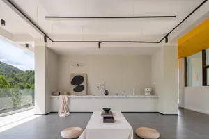 A minimalist modern living room with concrete floor and walls, featuring a white table and two wicker stools.