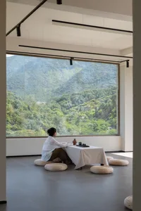 A modern residential interior featuring a cozy living space with white curtains on a large window, overlooking a serene outdo