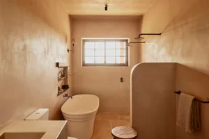 A minimalist bathroom with concrete walls, featuring a freestanding bathtub, a toilet, and a sink with a wooden stool. The ba
