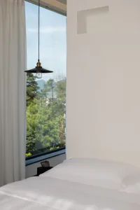 A modern white bedroom with a large window overlooking green trees and mountains, featuring a minimalist design with white be