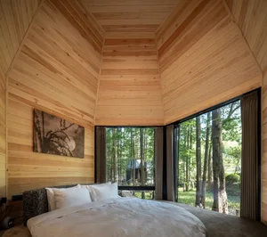 A minimalist bedroom featuring wood-paneled walls and a large window overlooking a forest.