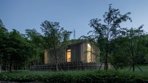 A minimalist building constructed primarily of wood, featuring a single illuminated window.