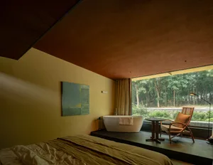 Modern bedroom featuring a ceramic armchair, a large window with a view of a forest, and a freestanding bathtub.
