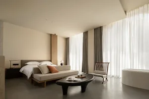 A modern minimalist bedroom featuring a bed with white bedding, neutral-colored cushions, and a sleek black coffee table.