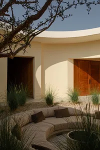 Modern outdoor lounge area with sand ground and greenery, featuring a modern sofa and cushions.