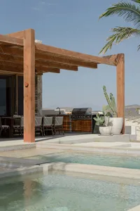 Modern residential pool area featuring a pergola-covered outdoor kitchen, grill, dining table, and cactus plants.