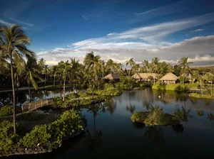 A serene waterscape featuring a resort with thatched-roof huts and lush vegetation under a clear, partly cloudy sky.