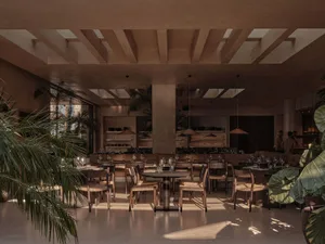 A modern restaurant dining room with wooden tables, glass pendant lights, and green plants.
