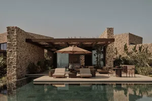 Modern residential exterior featuring a stone building, poolside lounge area, and wooden furniture.