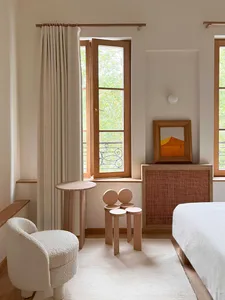 A minimalist bedroom with a bed, round side table, and chairs made of wood and textiles, featuring a window and artwork.