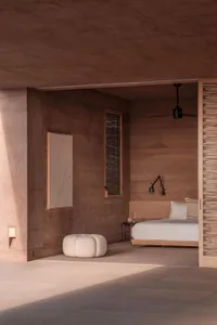 A minimalist bedroom with clay walls and a white bed, featuring a wall-mounted lamp, a window with view, and a pouf.