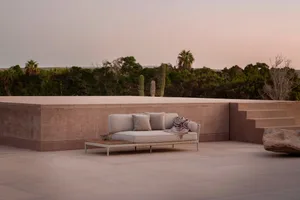 A minimalist outdoor lounge area featuring a concrete bench with cushions and a blanket, surrounded by natural vegetation.