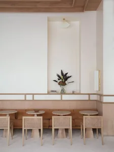 A modern cafe interior featuring a long wooden sofa, round tables, and cane-back chairs.