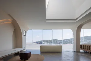 Modern living room with large glass windows overlooking the ocean, featuring sleek furniture and minimalist decor.