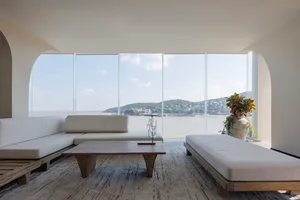 A modern living room featuring light wood and concrete furniture, large glass windows, and a view of a coastal landscape.