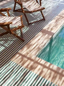 A sunny outdoor area featuring a tiled floor with striped patterns, wooden beach chairs, and a glimpse of a swimming pool.