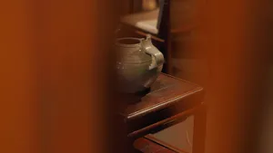A ceramic vase with a decorative design sits on a wooden shelf in a warmly lit room.