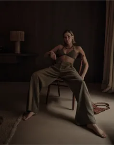 A woman sitting on a chair in a dimly lit room with brown walls, wearing a dark bikini top and loose pants.