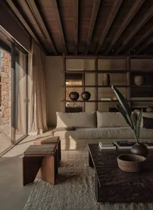 A modern, minimalist living room with wooden furniture, stone walls, and a large window allowing natural light to illuminate 