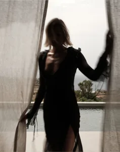 A person wearing a black dress standing by a window with sheer curtains, overlooking a pool and natural landscape.