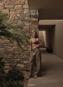 A woman wearing a minimalist outfit, including a bikini top and wide-leg pants, stands near a stone wall.