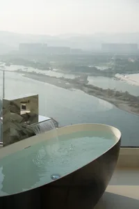 Modern bathroom with glass waterfall bathtub overlooking scenic river and cityscape.