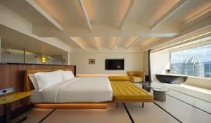 A modern hotel bedroom featuring a king-sized bed with a yellow leather headboard, a yellow leather daybed, and a large windo