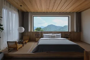 A modern bedroom with a wooden platform, large window overlooking mountains, white bedding, and minimal furniture.