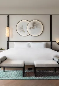A minimalist bedroom featuring a bed with white textiles, mounted artworks on the wall, and a wooden floor with metal accents