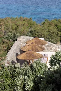 Three thatched umbrellas and sun loungers on a stone terrace overlooking the ocean and greenery.