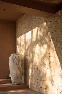 A close-up view of a corner featuring a textured stone wall with a large, weathered vase on a step, casting shadows and illum