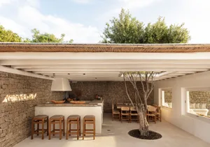 An outdoor kitchen area featuring a modern design with concrete countertops, stone walls, and timber stools and table.