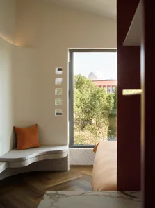 A minimalist bedroom featuring a concrete bench with a cushion and a window overlooking trees and a building.