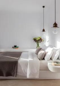 A minimalist bedroom with a bed covered in white linens and gray throw blanket, accompanied by a white side table, pendant li