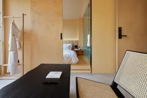 A minimalist bedroom with clay furnishings and light-colored walls, featuring a bed, desk, and chair.