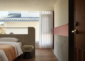 Minimalist bedroom with concrete and wood finishes, white bed, sheer curtains, and a view of a tiled roof.