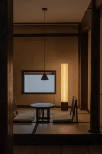 A minimalist living room with tan tones, featuring a small round table, cushioned floor seating, a tall cylindrical lamp, and