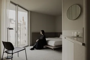 A minimalist bedroom with white furniture and a wooden bed, featuring a large window and a black chair.