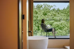 A modern interior featuring a large window with a view of green trees, a white bathtub, and an armchair with a person sitting