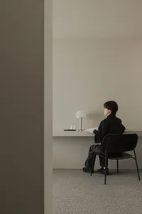 A minimalist home workspace with a concrete desk, black chair, and a white lamp on the desk.
