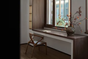 A modern library space featuring a wooden desk, chair, and bookshelves with a vase of autumn leaves on the windowsill.
